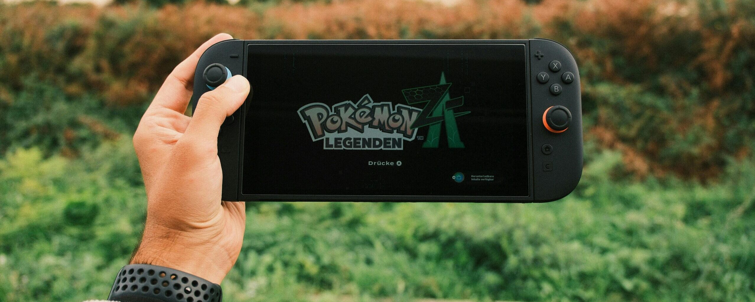A person holds a handheld gaming device outdoors with Pokémon Legends on screen.